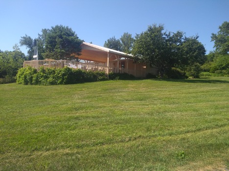 View of the deck from the east. The deck is surrounded by grass in the foreground, and lined by bushes