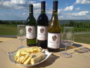 A bottle of Seyval, Chambourcin, and Chardonnay set between two Frontenac Point glasses and a cheese plate on a sunny day winery deck with Cayuga Lake in the background. 