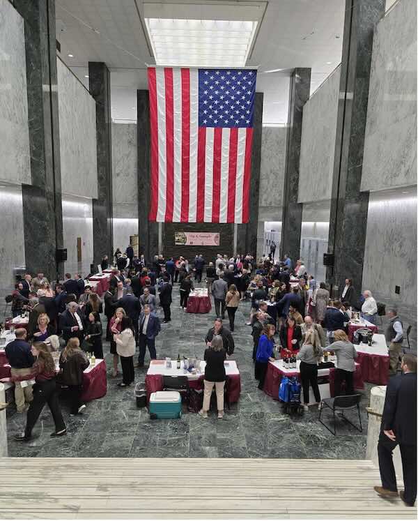 Winery owners serve wine samples in an exhibition room with a large American flag hanging in the center of the back of the room